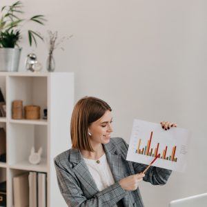 Young business woman in white office with smile shows graphic, talking to customers on Skype